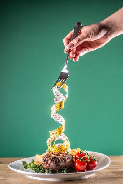 Hand with fork swirling measuring tape from cooked steak with vegetables on a plate