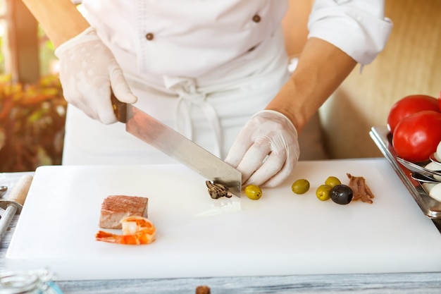 Hand with knife cuts olive. Shrimp on white cooking board. Chef working at the table. Work is in full swing.