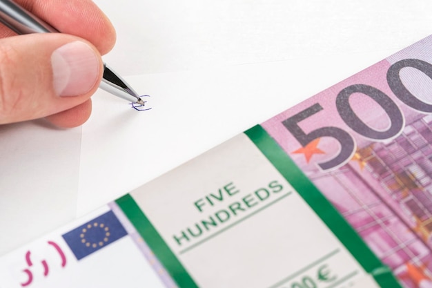 Hand with pen writes euro sign on white sheet, next to a bundle of money