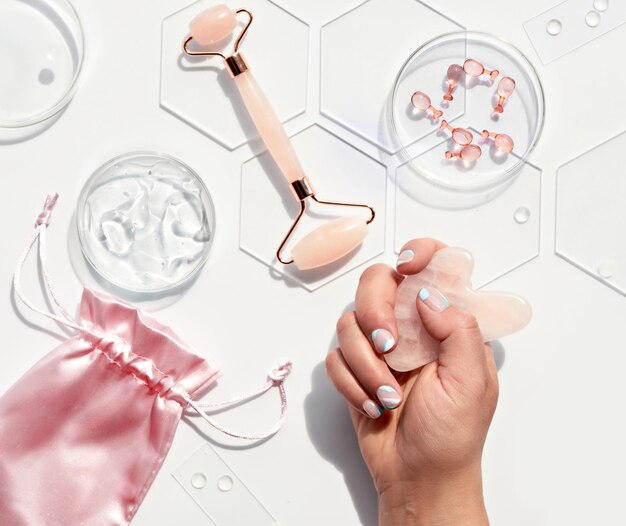 Hand with pink quarts face roller, gua sha stone, serum capsules in chemical glass dish, hexagons. Natural cosmetics laboratory. Off white flat lay with pink beauty objects, monochromatic look.