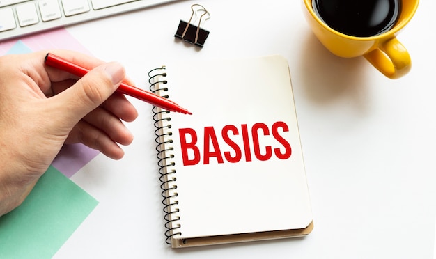 Hand with red pen, Cofee cup, Stick, Keyboard and white notebook with text BASICS