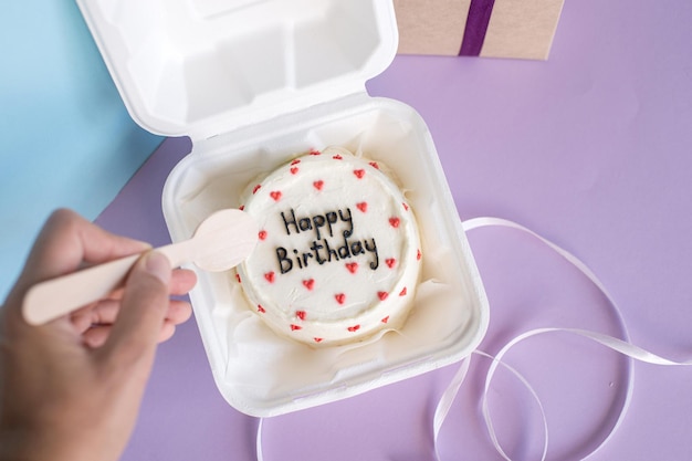 A hand with a wooden spoon reaches for a festive bento cake near the gift box