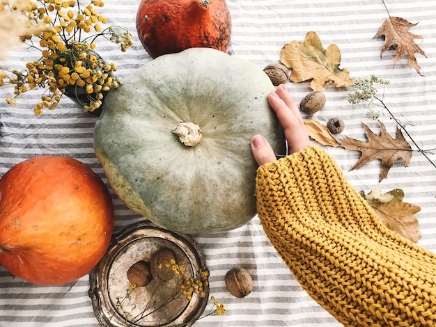 Photo hand in yellow sweater holding pumpkin and nuts yellow flowers fall leaves on rustic table flat lay fall decor and arrangement on table autumn harvest thanksgiving concept