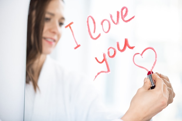 Photo hand of young amorous brunette female writing love message and drawing small heart with red or crimson lipstick on mirror