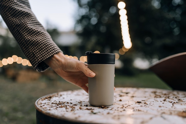 Photo the hand of a young girl in a stylish coat takes a thermos cup with a hot drink outdoors