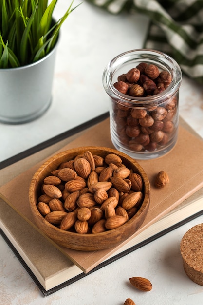 a handful of almonds in a wooden plate and hazelnuts in a glass jar on a light background and a green plant in the back the background