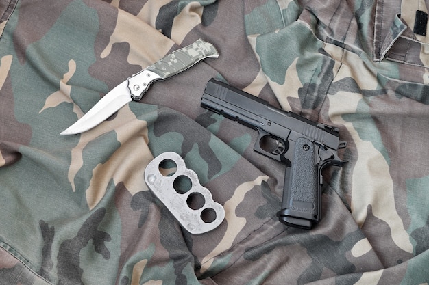 Handgun lies with brass knuckles and knife on camouflage military uniform close up. Concept of looting and illegal arms trade