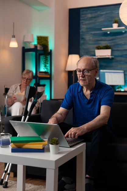 Handicap retired people relaxing at modern home with technology and medical equipment. Old man using digital laptop computer device sitting on sofa with crutches and woman in wheelchair