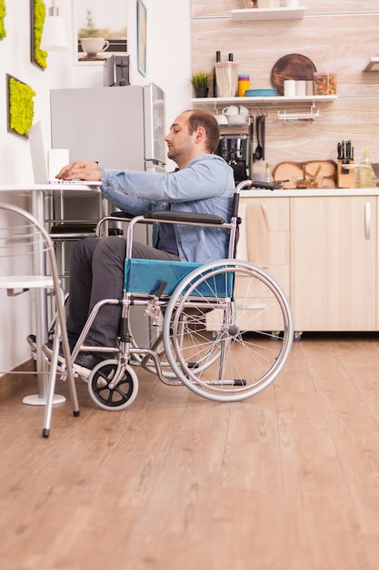 Handicapped businessman in wheelchair using laptop in kitchen. Disabled paralyzed handicapped man with walking disability integrating after an accident.