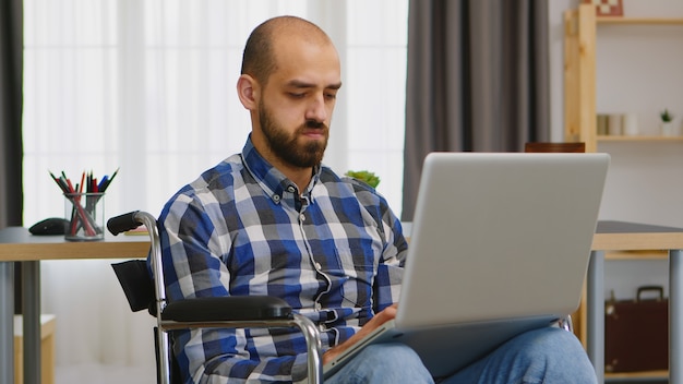 Handicapped freelancer in living room in wheelchair working on laptop .