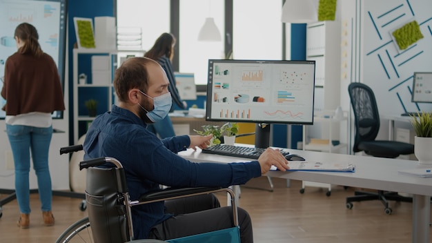 Handicapped invalid paralyzed man and teamworkers working in new normal office at financial strategy wearing protective mask