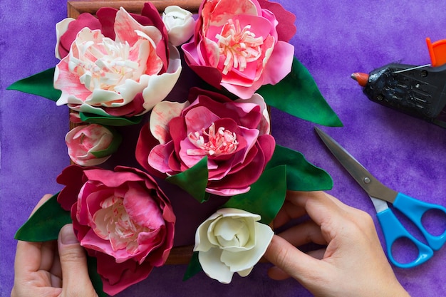 Handmade decorative peonies made from foamiran on wooden panel holding by woman hands and glue gun and scissors