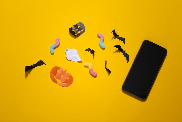 Handmade Halloween decor and gummy worms, smartphone on yellow background. Halloween background. Top view. Flat lay