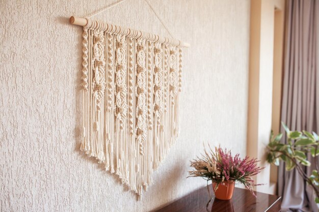 Photo handmade macrame wall decoration with wooden stick hanging on a white wall female hobby eco friendly modern knitting natural decoration in interior copy space