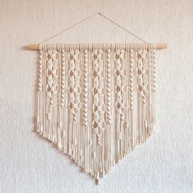 Photo handmade macrame wall decoration with wooden stick hanging on a white wall female hobby eco friendly modern knitting natural decoration in interior copy space