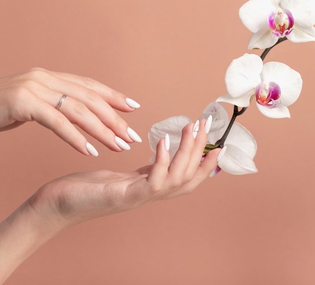 Hands of a beautiful wellgroomed woman with feminine nails on a beige background with orchid flowers Manicure pedicure beauty salon concept On nails white gel polish with an abstract pattern