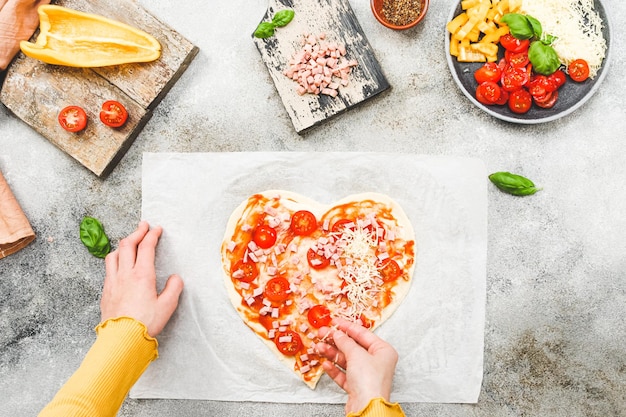 Hands of caucasian teenage girl lay down sliced cherry tomato on heartshaped pizza dough