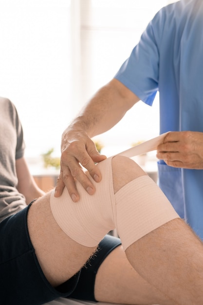 Hands of clinician in blue uniform wrapping injured knee of young patient with flexible bandage before rehabilitation exercises in medical center