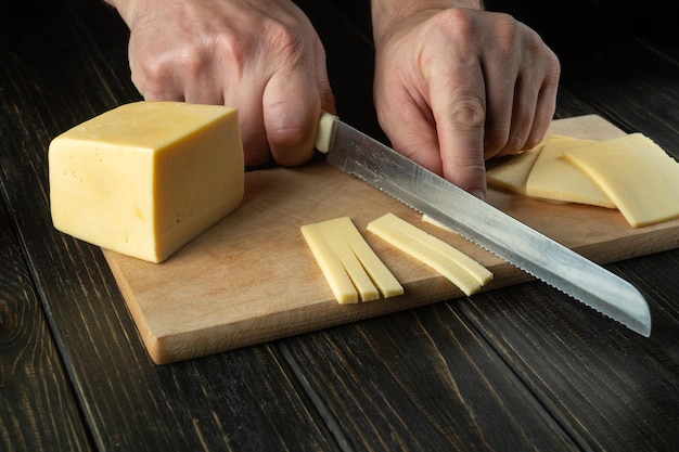 The hands of the cook with a knife cut the cheese into small pieces for tasting on a cutting board