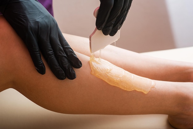 Hands of a cosmetologist in black gloves applying sugar paste with spatula on a woman's leg Depilation procedure