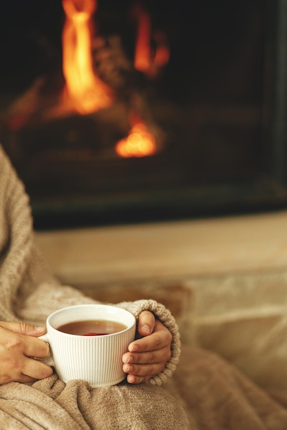 Hands in cozy sweater holding cup of warm tea on background of burning fireplace close up hygge