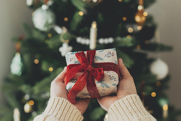 Hands in cozy sweater holding stylish christmas gift in festive wrapping paper with bow on background of christmas tree lights Happy holidays Atmospheric christmas time