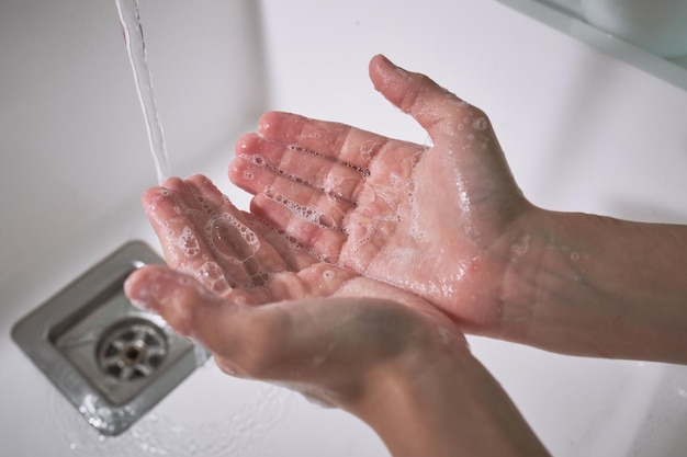 Hands of crop faceless kid washing hands with foamy soap in sink with water stream during hygienic routine in bathroom