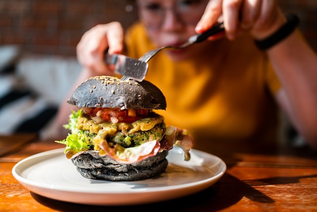 Hands cuts Broccoli quinoa charcoal burger topped with guacamole, mango salsa and fresh salad by knife and fork, served in a white plate. Creative vegan meal for vegetarians.