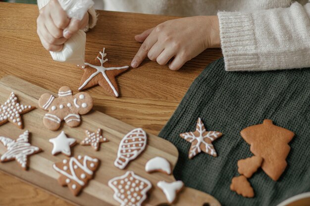 Photo hands decorating christmas gingerbread cookies with icing on rustic wooden table top view atmospheric christmas holiday traditions decorating baked cookies with sugar frosting family time