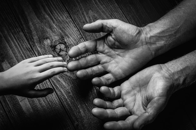 Hands of an elderly man holding the hand of a younger man. Lots of texture and character in the old man hands. on black wooden wall.black and white.