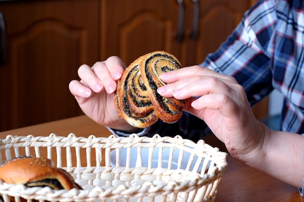 The hands of an elderly woman operate a sweet bun with poppy seeds in the kitchen at a brown table without a face closeup