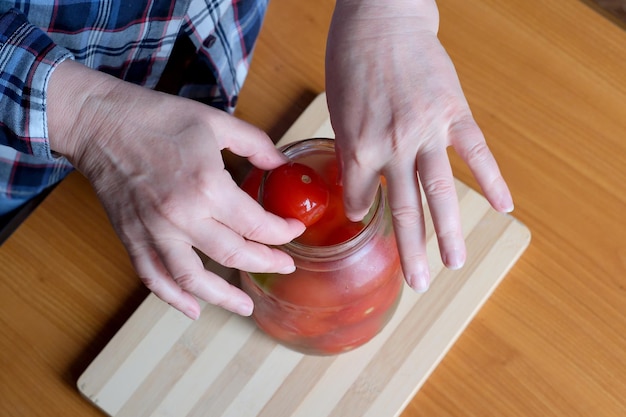 The hands of an elderly woman take out delicious homemade pickled tomatoes from a jar in the kitchen at a brown table without a face closeup