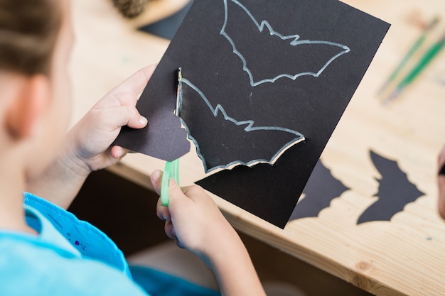 Photo hands of elementrary pupil cutting out halloween bat from black paper while sitting by wooden desk at lesson