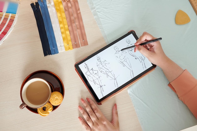 Hands of female designer drinking cup of coffee with cookies and drawing fashion sketch on digital t...