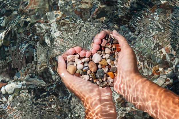 Hands full of colorful pebbles forming heart shape on the sea waves background. Holiday, rest, vacation at the seaside concept