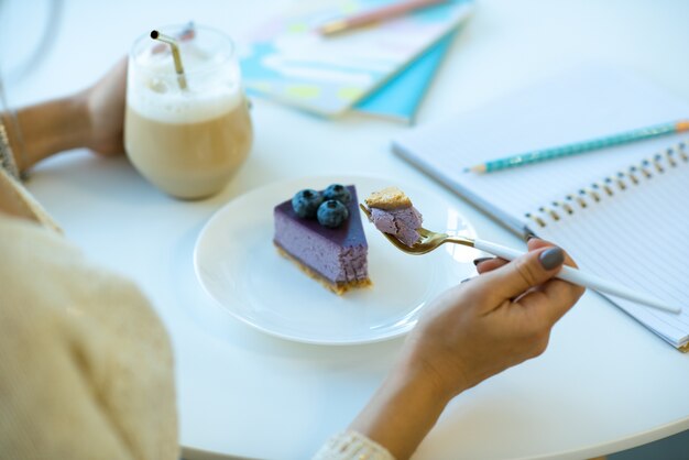 Photo hands of girl holding fork with piece of tasty blueberry cheesecake over plate while enjoying dessert with cappuccino in cafe