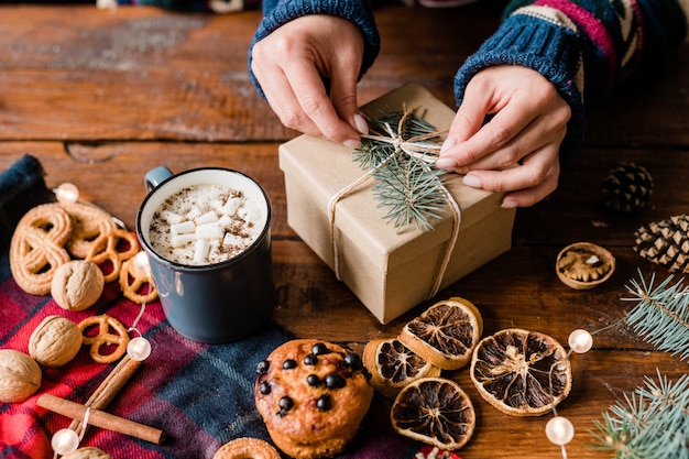 Hands of girl making knot on top of wrapped giftbox surrounded by sweet food, hot drink in mug and walnuts