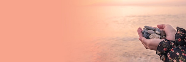 Hands hold large pebbles in their palms against the backdrop of calm sea water in the light of the setting sun