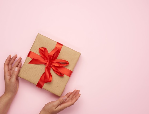 Hands hold a square box wrapped in brown paper and tied with a red silk ribbon