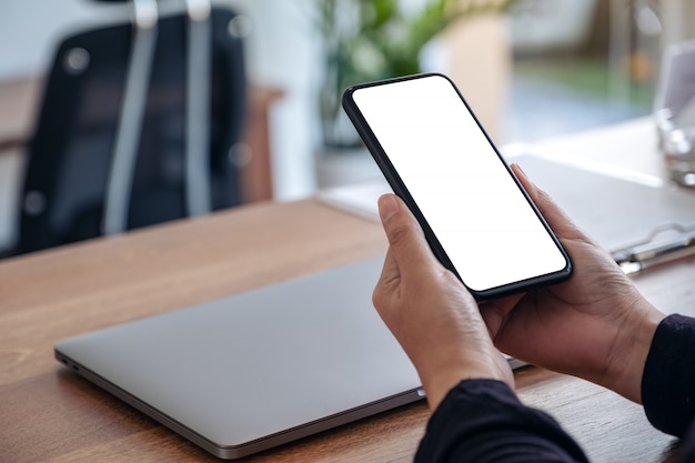  hands holding black mobile phone with blank white screen with laptop on wooden table