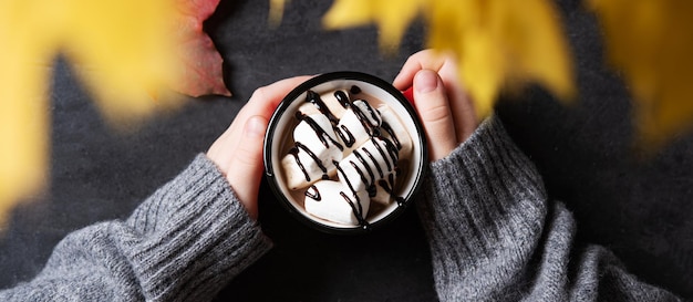 Hands holding a cup of hot chocolate with marshmallow on a dark gray background Autumn concept of a warming drink Top view Horizontal orientation