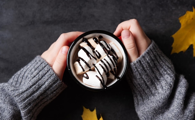 Hands holding a cup of hot chocolate with marshmallow on a dark gray background Autumn concept of a warming drink Top view