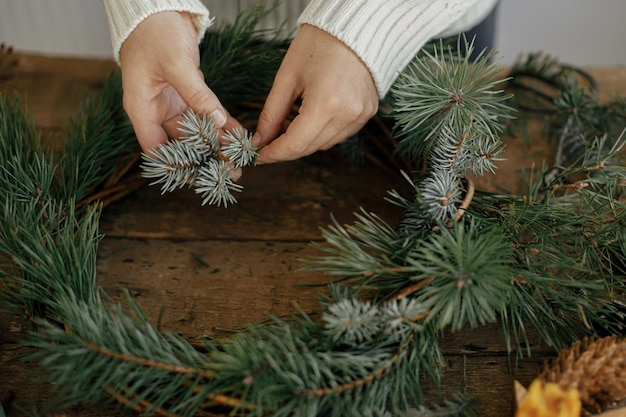 Hands holding fir branch and arranging christmas wreath on rustic background Winter Workshop