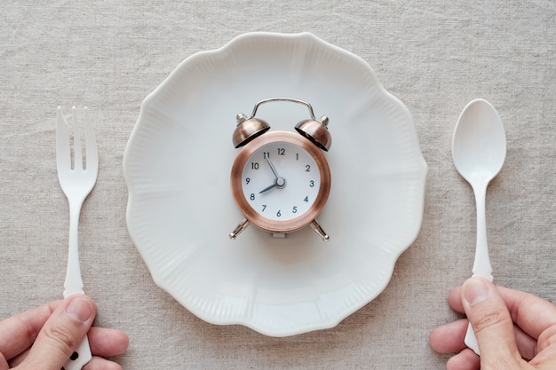 Photo hands holding fork and spoon and a clock on the plate, intermittent fasting diet concept