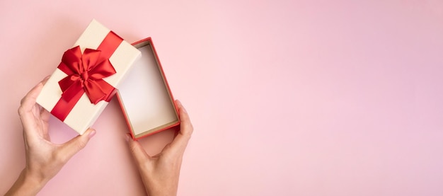 Hands holding open gift box with red bow on pink background Top view