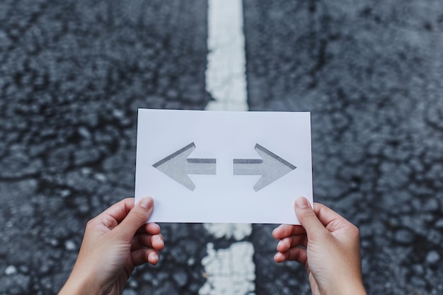 Photo hands holding paper with arrows crossroad symbol splitted in three different directions choose the correct way between left right and front difficult decision concept over asphalt road background