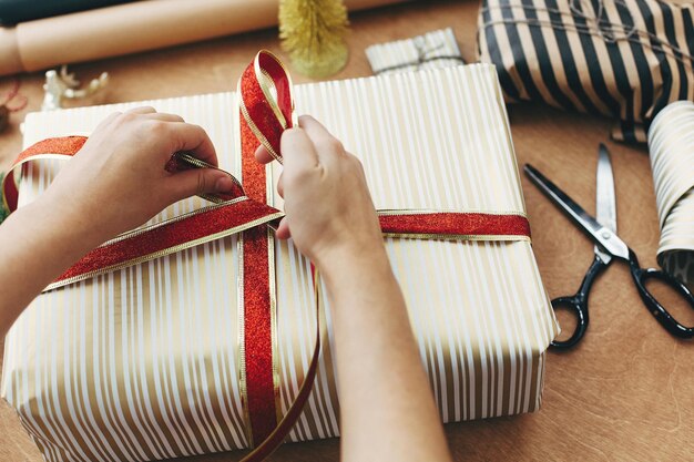 Hands holding red ribbon and wrapping stylish gift box in striped golden paper and scissors presents golden tree deer on wooden table Wrapping christmas gifts concept