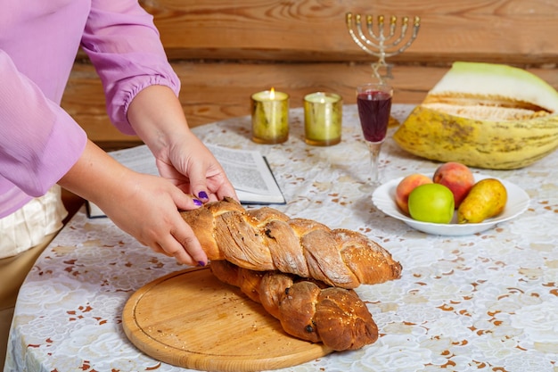 The hands of a Jewish woman break the challah after the blessing during the Shabbat meal next to burning candles and a glass of wine