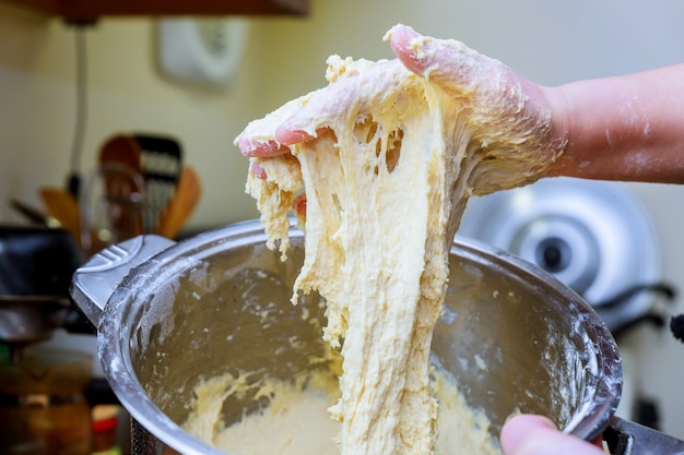 hands knead dough on table, surrounded with much flour, bowls, eggs and milk. 