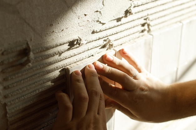 Hands laying modern square tile on adhesive close up Worker installing stylish white tiles on plaster wall in sunlight Renovation